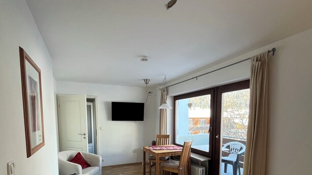 Una habitación de hotel con cama, una mesa de comedor con sillas, un televisor montado en la pared y un ventanal con cortinas.