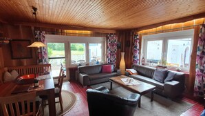 A cozy room with a wooden ceiling, a sofa, a dining table with chairs, and large windows with floral curtains.