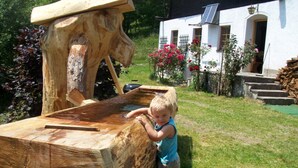 Niño interactuando con una gran escultura de madera al aire libre, con una casa y jardín de fondo.