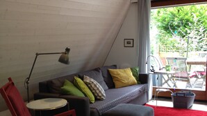 Una habitación acogedora con techo inclinado, un sofá gris con cojines coloridos, un sillón mecedor rojo, una mesita redonda y una alfombra roja.