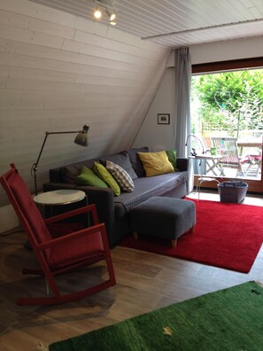 Une pièce chaleureuse avec un plafond en pente, un canapé gris agrémenté de coussins colorés, un fauteuil à bascule rouge, une petite table ronde et un tapis rouge.
