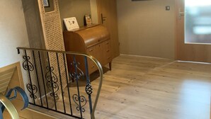 Un escalier en bois, avec une rampe en métal, mène à un palier. Sur ce palier, on trouve une commode en bois et un tableau encadré.