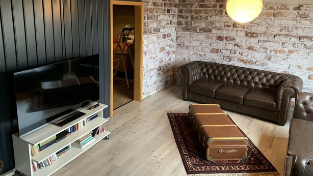 Un salon moderne avec un mur lambrissé en bois foncé, un mur en briques, un canapé en cuir, un téléviseur, une bibliothèque et une valise posée sur un tapis.