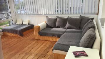 Ein gemütliches Zimmer mit einem Korbssessel, einem kleineren Sofa und einem weißen Tisch mit einem Buch.
