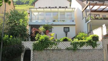 Ein zweistöckiges Haus mit Balkon, umgeben von einer Steinmauer und verschiedenen Pflanzen.