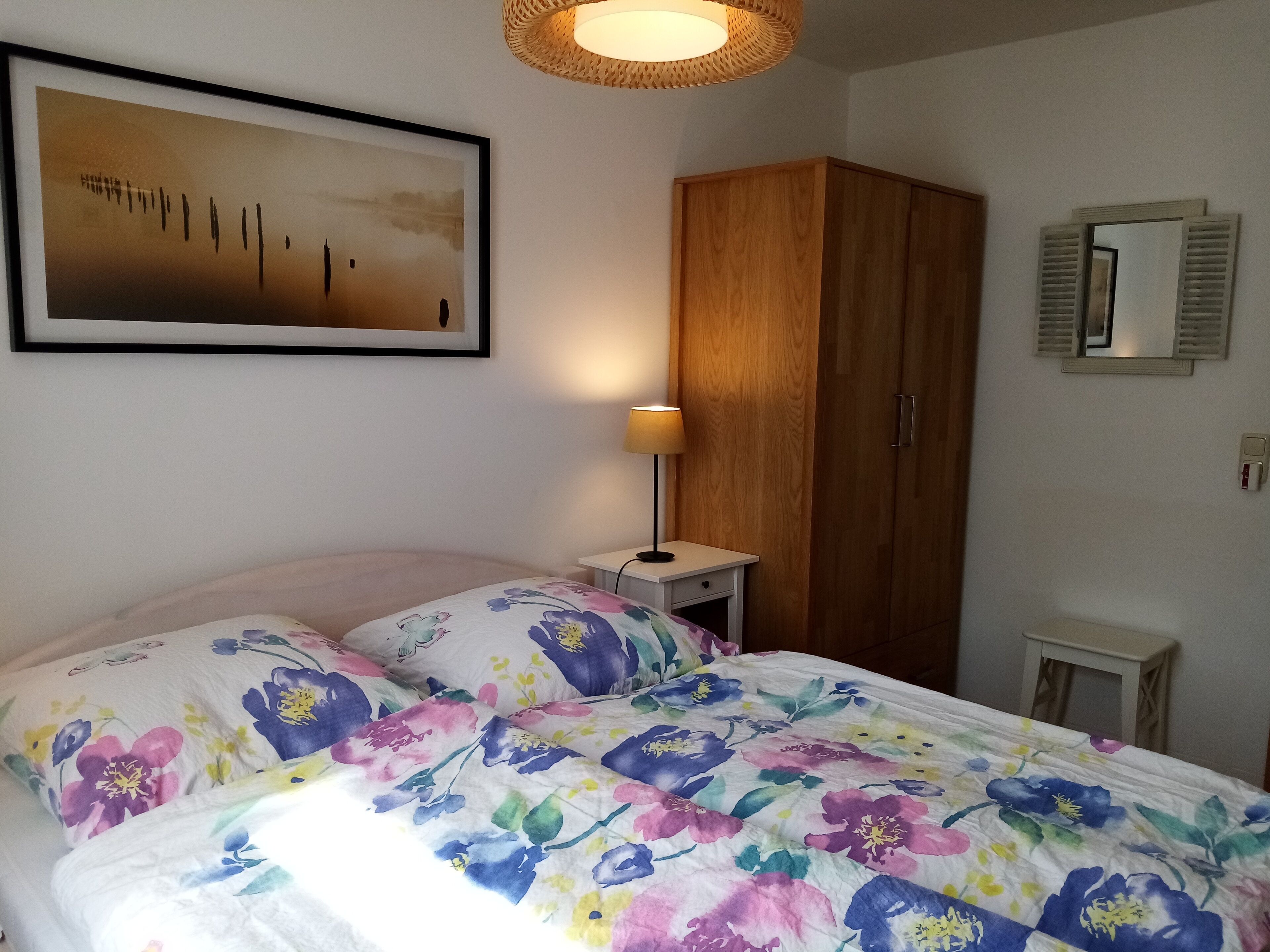 Une chambre à coucher comprenant un lit, une table de chevet, une armoire en bois, un miroir fixé au mur et un tableau encadré.