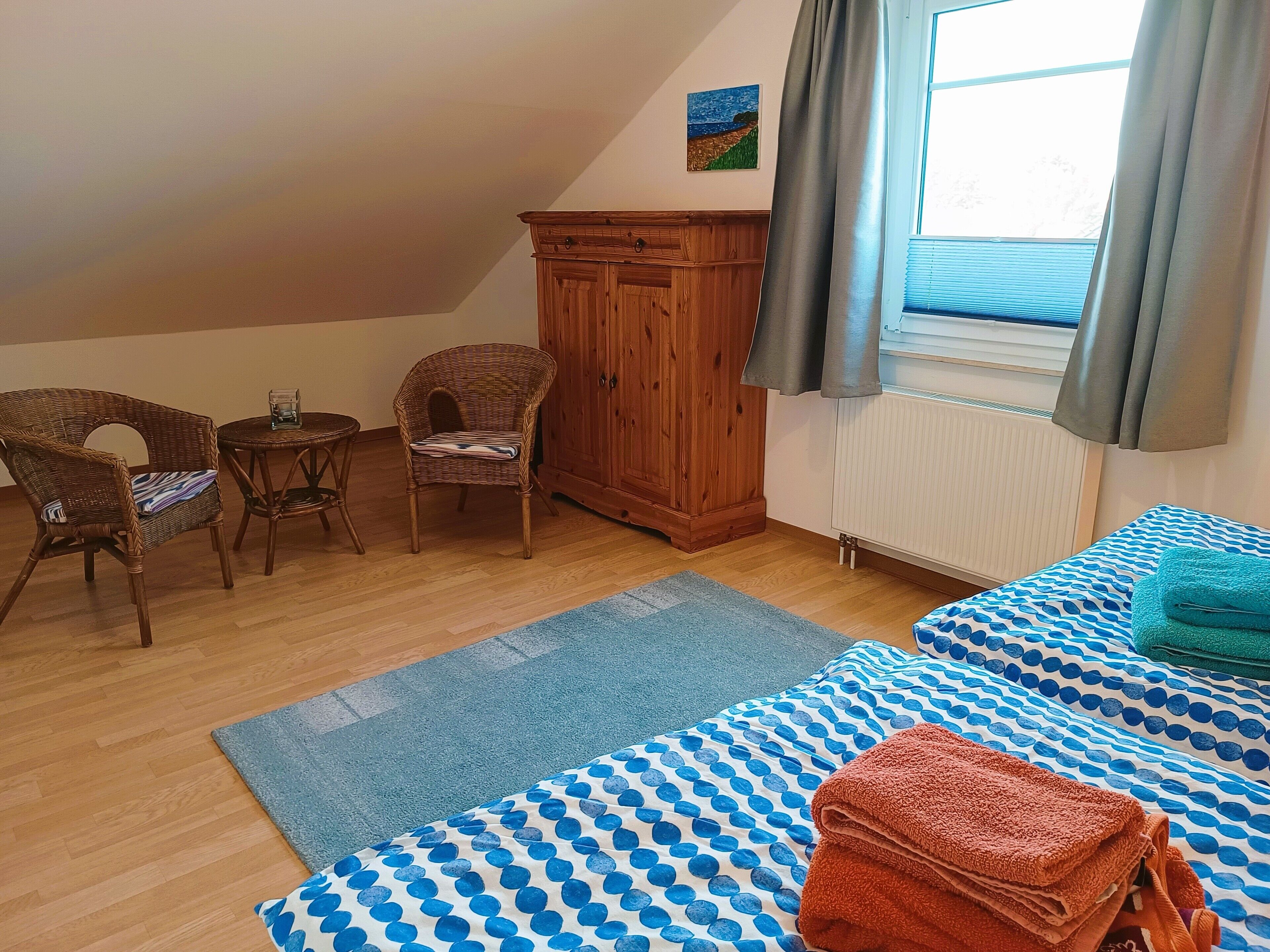 A cozy room with two beds, a wooden wardrobe, a wicker chair, a small table, and a window with curtains.