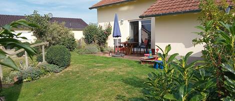 Une maison avec un toit en tuiles, une terrasse avec une table et des chaises, et un jardin avec différentes plantes.