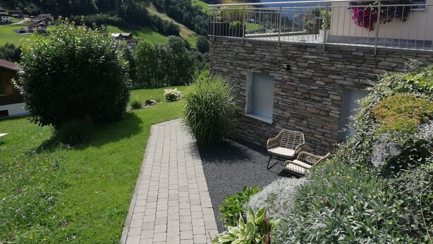 Un edificio de piedra con balcón, un camino empedrado y un jardín con diversas plantas y una silla.