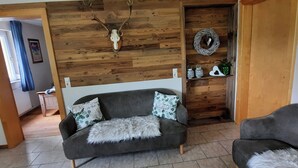 Un salón con un sofá gris, una manta mullida, una pared de madera con un cráneo de venado montado, una corona de mimbre y una estantería con objetos decorativos.