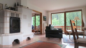 A living room with a fireplace, a sofa, a dining table, and large windows offering a view of the outdoors. - In the southern Black Forest. Pure nature, close to the lake, family-friendly (Baden-Württemberg)