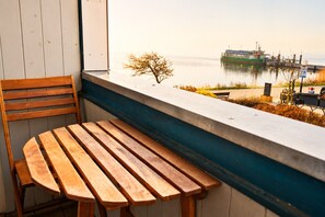Un banc et une chaise en bois, placés près d’une fenêtre donnant sur un plan d’eau où se trouvent des bateaux.