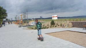 Un camino pavimentado con una persona practicando skate, un muro de ladrillo y una zona verde con un banco.