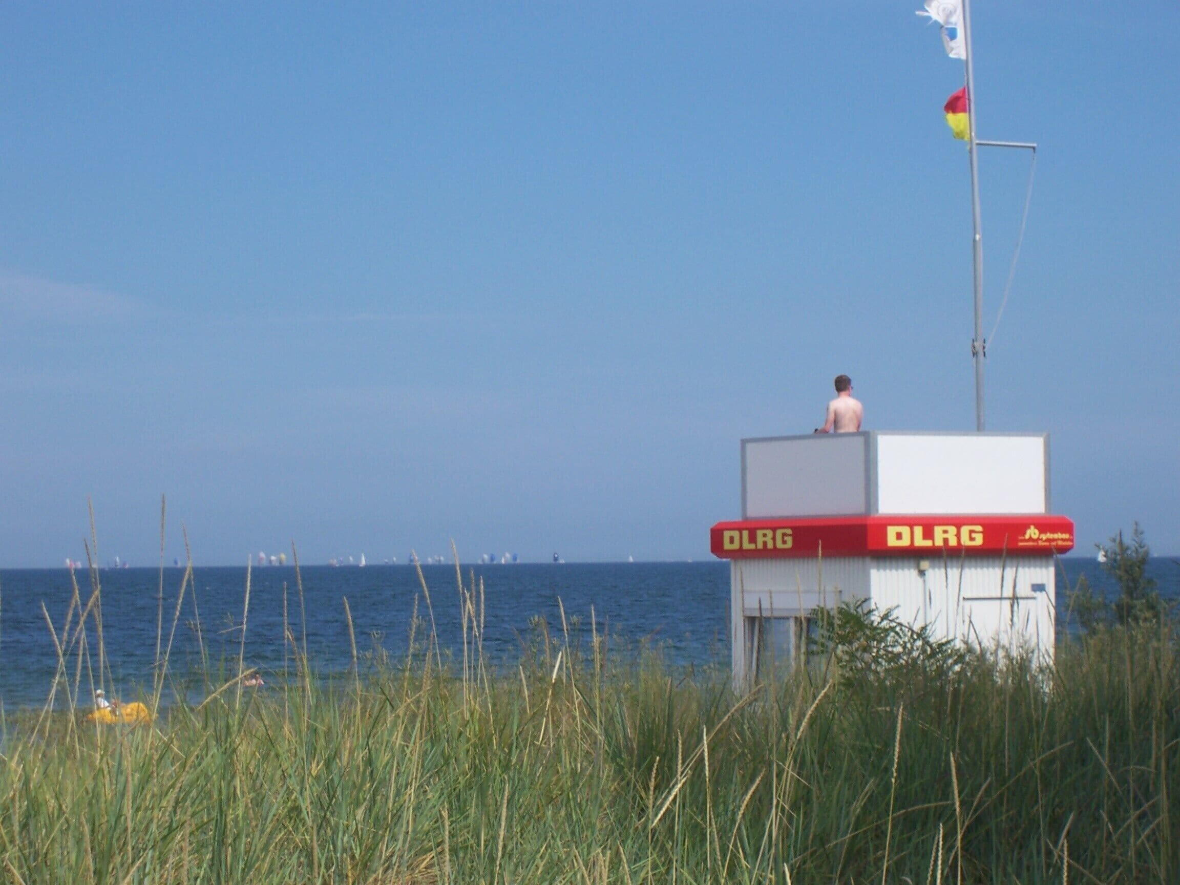 Ein Rettungsschwimmturm mit dem Schriftzug 'DLRG', der auf ein Gewässer mit Booten blickt.
