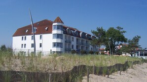 Un hotel de varios pisos con techo de tejas rojas, paredes exteriores blancas y balcones. Se encuentra cerca de una playa arenosa con vegetación escasa y una valla de madera.