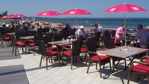 Un área de comedor al aire libre con mesas y sillas, cojines rojos y sombrillas rojas.