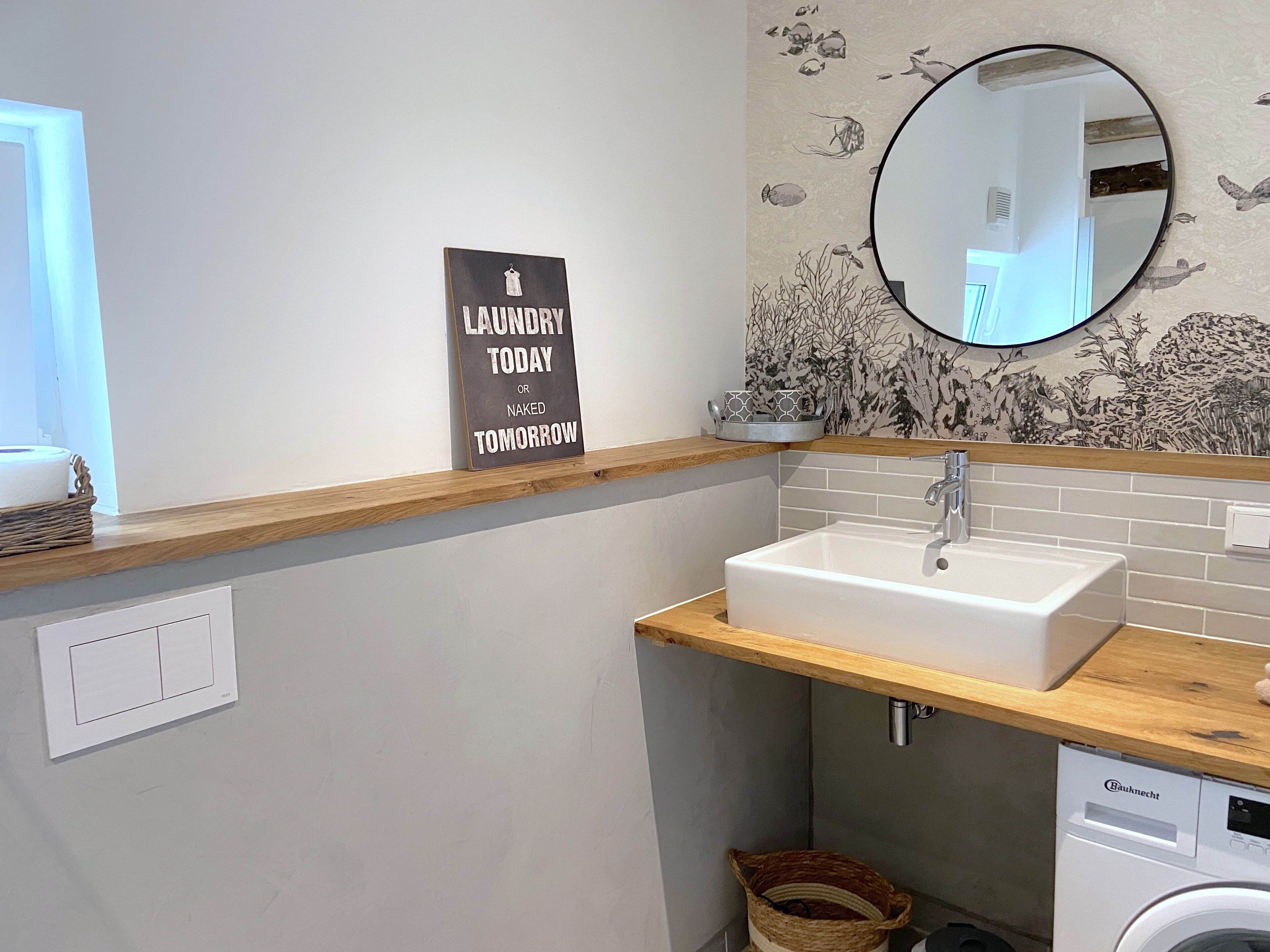 A modern bathroom with a wooden countertop, a white sink, a Bauknecht washing machine, and a sign that reads 'Laundry Today or Naked Tomorrow'.