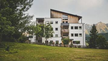 Ein modernes, mehrgeschossiges Gebäude mit flachem Dach und großen Fenstern, umgeben von Grünflächen und Bergen im Hintergrund.