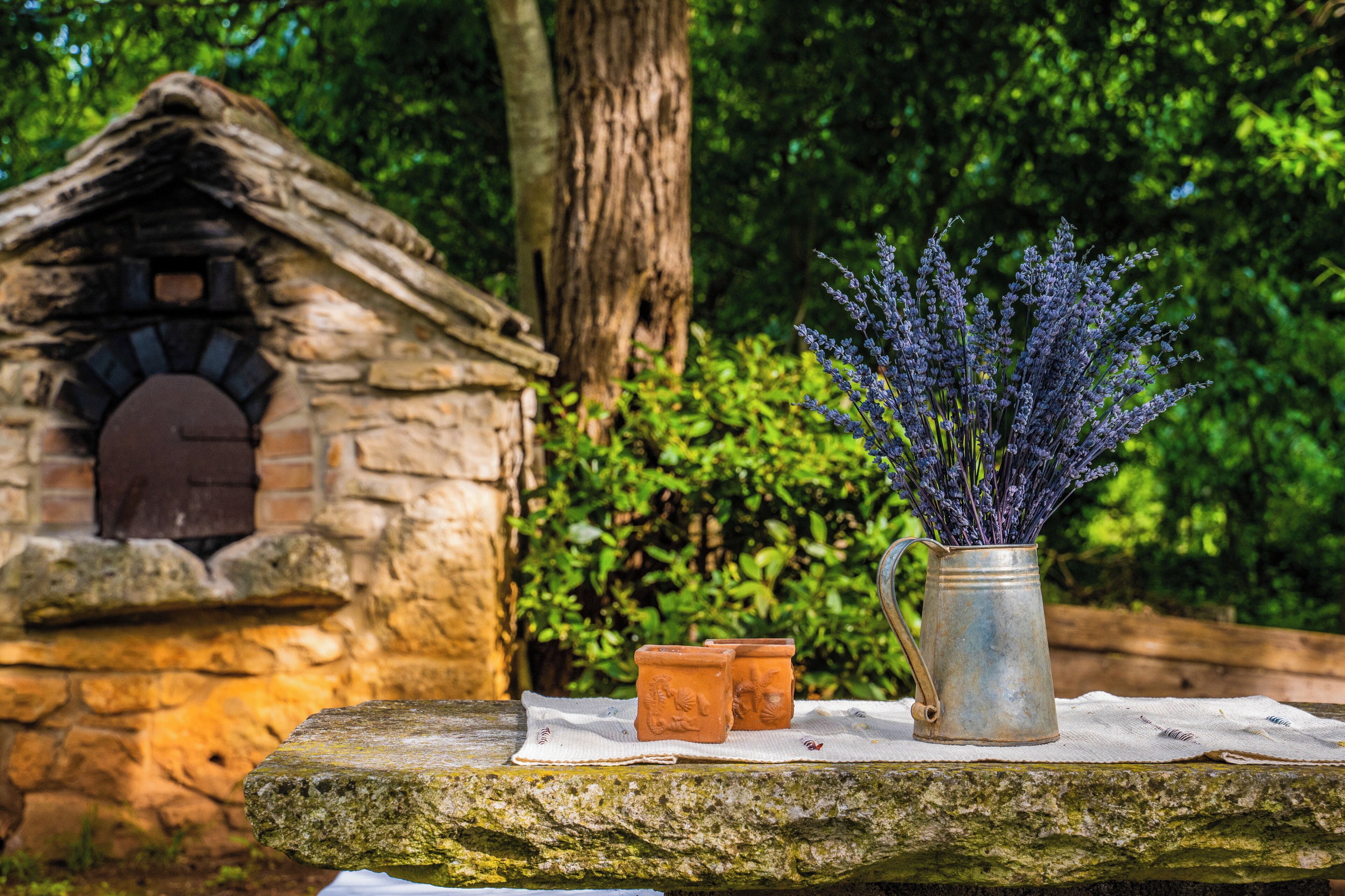 A rustic stone house with a thatched roof, a metal vase with lavender, and terracotta pots on a stone ledge.