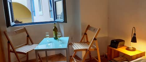 A small room with a glass table, two wooden chairs, a bottle of wine, two glasses, a lamp, and a bed with a striped blanket.