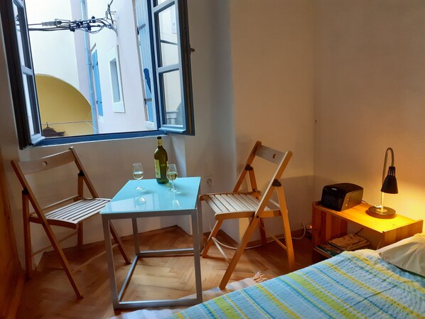 A small room with a glass table, two wooden chairs, a bottle of wine, two glasses, a lamp, and a bed with a striped blanket.