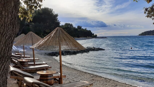 Una spiaggia con lettini di legno e ombrelloni di paglia, un litorale roccioso e una vista limpida sul mare.