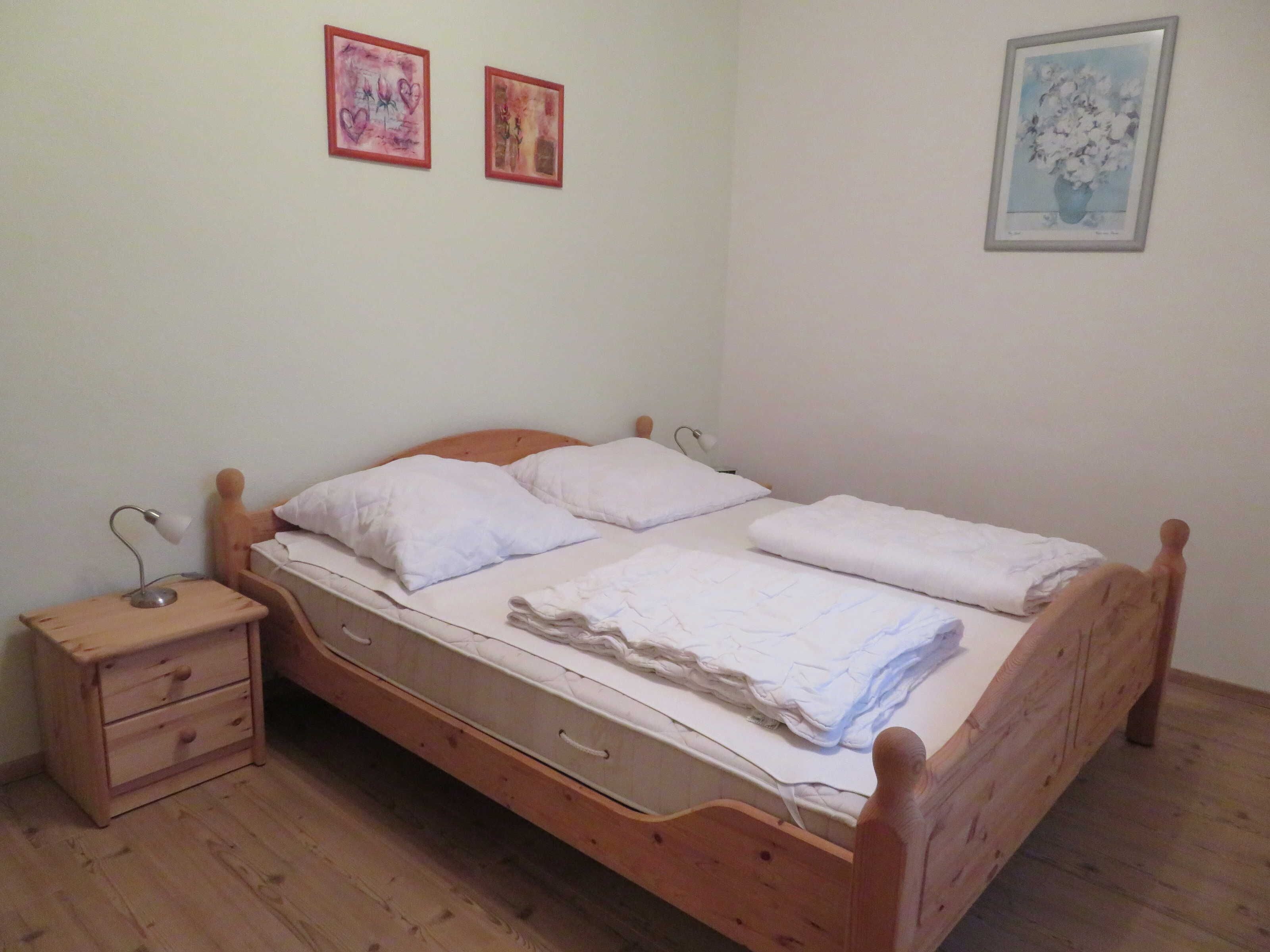 Ein Zimmer mit einem Einzelbett, einem Holzkopfstück, einem Nachttisch mit Schubladen und einer Nachttischlampe. An der Wand hängen zwei gerahmte Bilder.