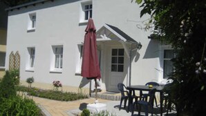 A white building with a patio area, a table with chairs, and a red umbrella. - Jasmund - Pension Waldrose (Sellin)
