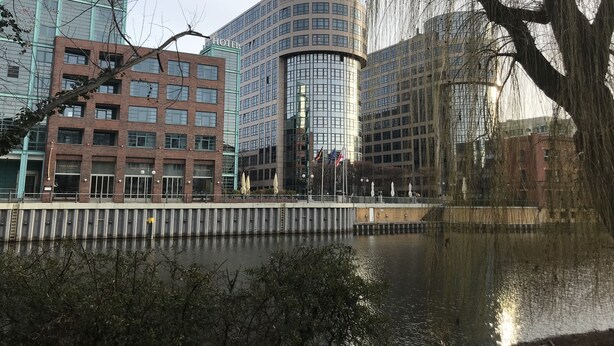 A modern hotel with a curved glass facade, a brick building with a Carl's Bakery sign, and a reflective water body in the foreground. - Details about our apartment. (Berlin)