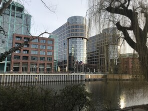 Een modern hotel met een gebogen glazen gevel, een bakstenen gebouw met een bord van Carl's Bakery, en een reflecterend wateroppervlak op de voorgrond.
