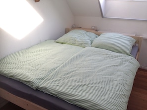 Een eenpersoonsbed met een groen-wit gestreept laken en kussens, een houten hoofdbord, en een raam boven het bed.