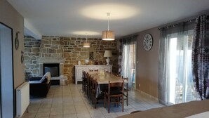 Amplia sala de estar con chimenea de piedra, una mesa de comedor con sillas, un sofá y una zona de cocina con estufa e iluminación colgante.