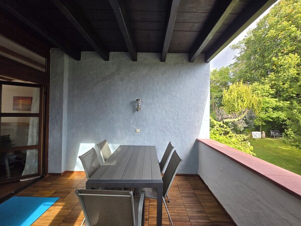 An outdoor patio with a table and chairs, a wooden ceiling, and a view of greenery.