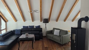 Ein Loft-Wohnzimmer mit freiliegenden Holzbalken, einem schwarzen Ledersofa, einem grauen Sofa, einem Glastisch und einem schwarzen Kamin.