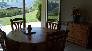 A dining area with a table and chairs, a vase with flowers, and a view of a garden and mountains through a glass door. - Charming apartment in chalet, close to ski slopes and town center    (Gérardmer)