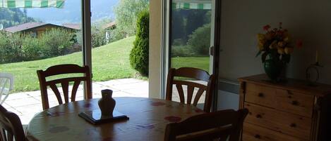 A dining area with a table and chairs, a vase with flowers, and a view of a garden and mountains through a glass door.