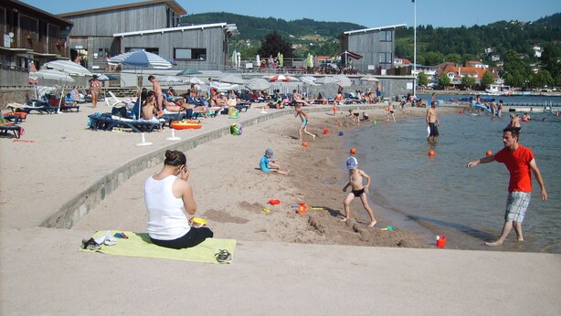 A beach with people sunbathing, swimming, and playing. There are umbrellas, lounge chairs, and a sandy area. - Charming apartment in chalet, close to ski slopes and town center    (Gérardmer)