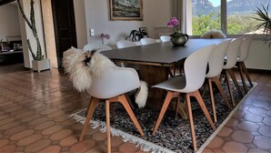 A dining area with a wooden table, white chairs, a rug, and a view of greenery through large windows.