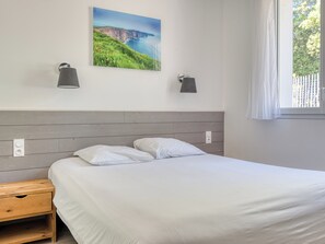 Una camera da letto con un letto, comodini, una lampada a parete e un quadro raffigurante un paesaggio costiero.