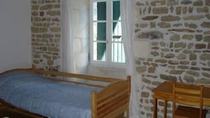 Una habitación con pared de piedra, una cama de madera, una ventana con cortinas y una pequeña mesa de madera.