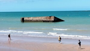 A beach with a concrete structure extending into the water, a clear sky, and three people walking along the shore.