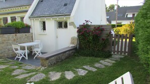 Ein kleines Haus mit weißer Fassade, einer Steinmauer, einer Holzterrasse und einem Garten mit trittfesten Pflastersteinen und Blumen.