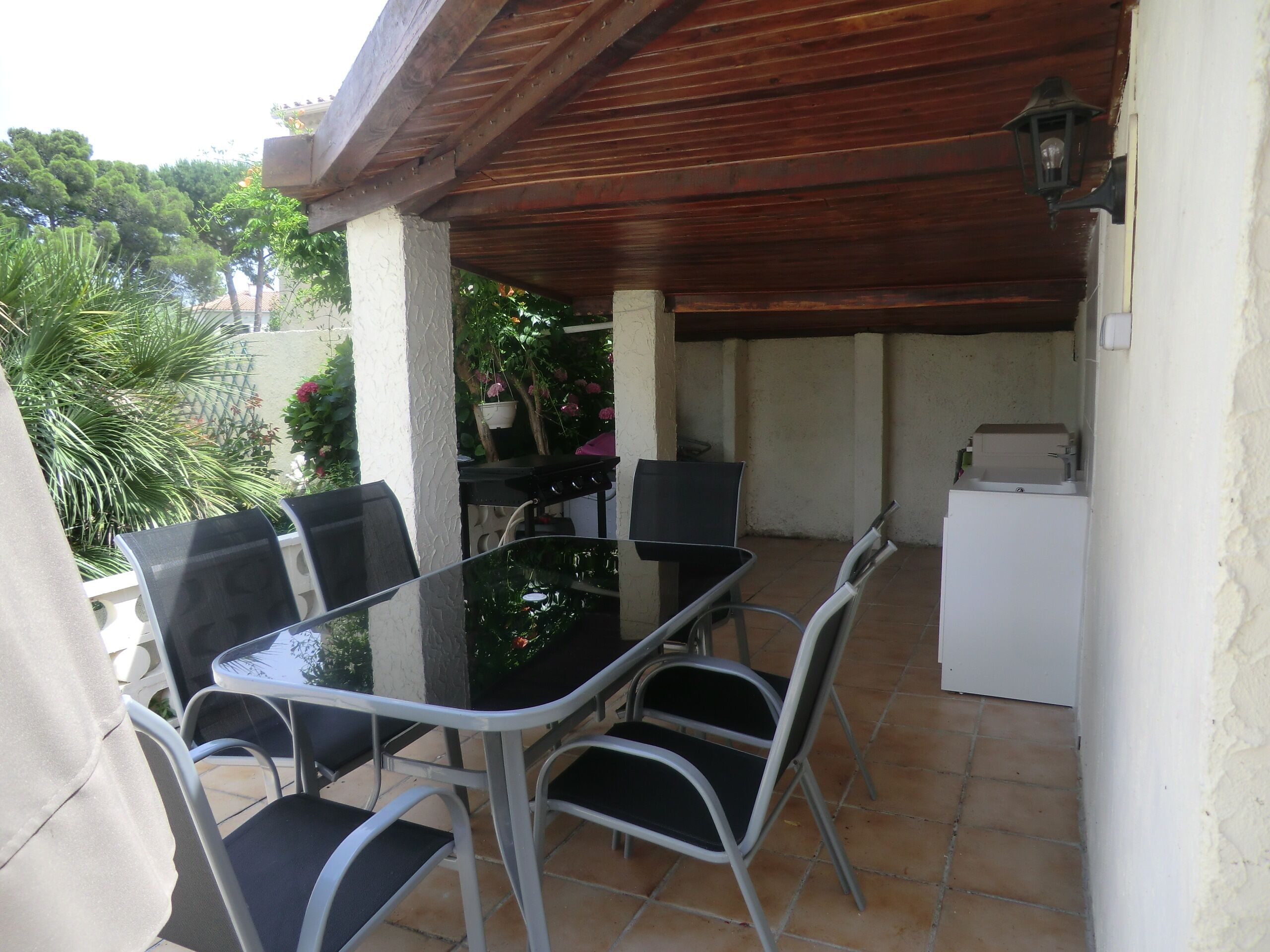 An outdoor patio with a glass table and four chairs, a small refrigerator, and a hanging planter with flowers.