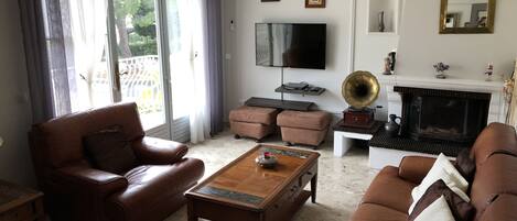 A living room with leather sofas, a wooden coffee table, a flat-screen TV, a fireplace, and a balcony with curtains.