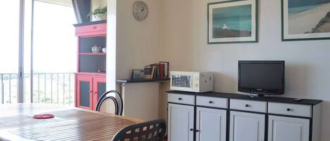 A compact living space with a dining table, chairs, a kitchenette with a microwave, a TV, and a small cabinet. There is a window with a view of the outdoors.