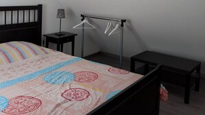 A bedroom with a sloped ceiling, a bed with a patterned bedspread, a bedside table with a lamp, a small table, and a clothing rack.