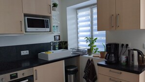 A compact kitchen with a microwave, sink, and stove. There is a window with blinds, a potted plant, and a kettle on the counter.