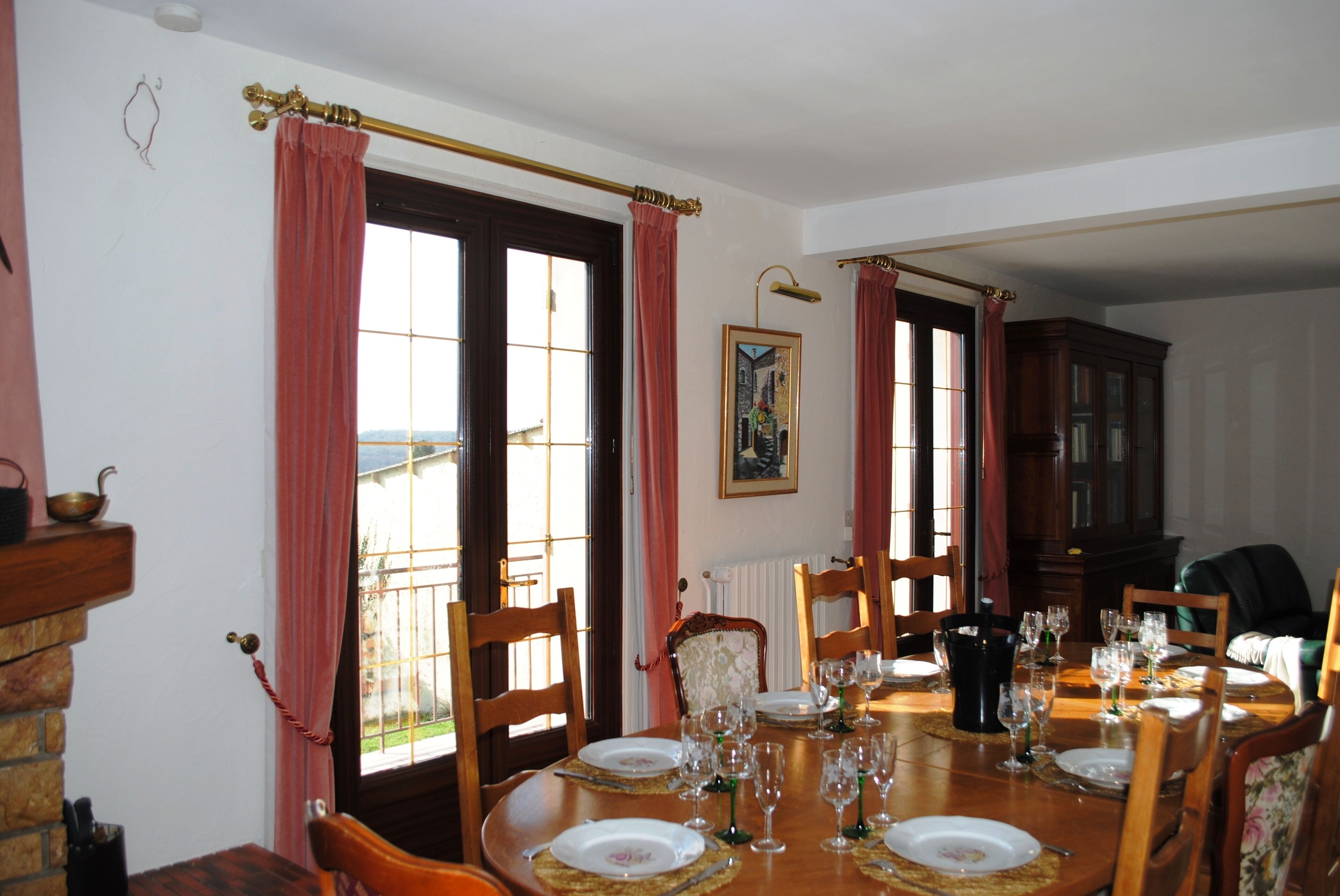 A dining room with a wooden table set for six, surrounded by chairs, a fireplace, and a view through the window.