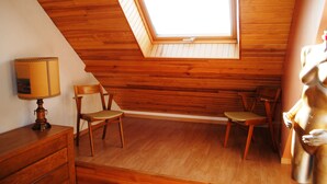 Een kamer met schuine wanden, houten lambrisering en een dakvenster. Er staan twee houten stoelen en een kleine tafel met een lamp.