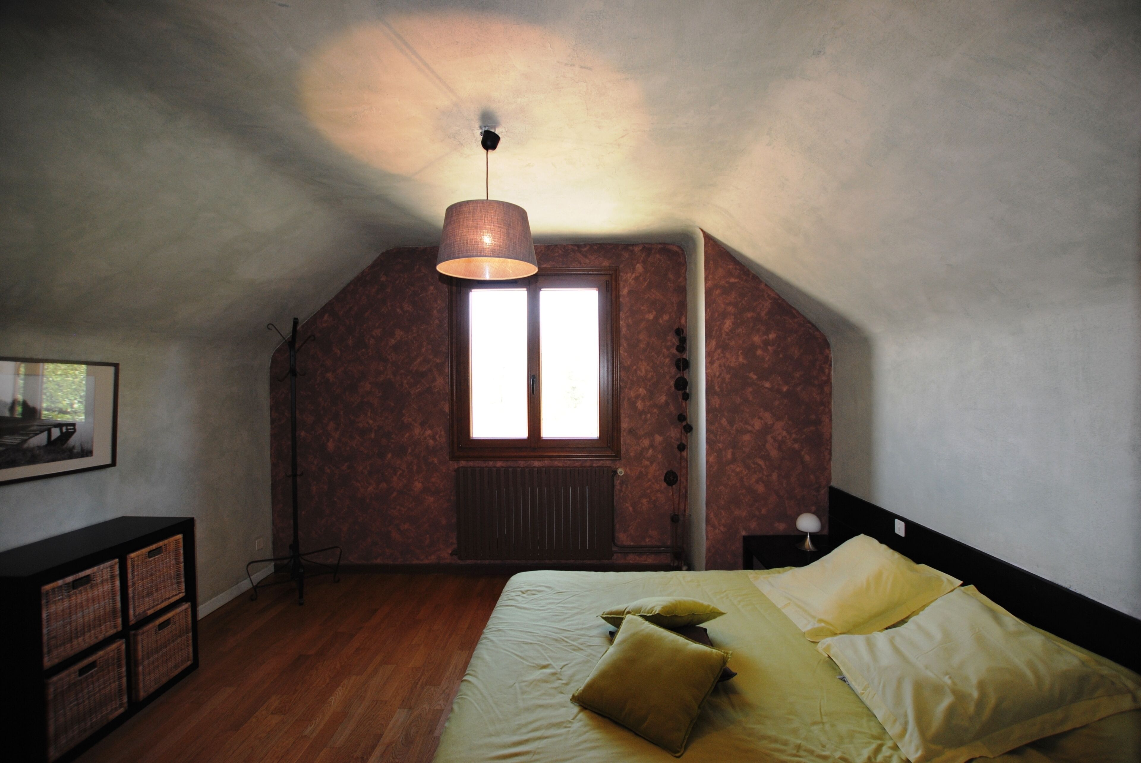 Ein Schlafzimmer mit schrägen Wänden, einem Fenster mit Heizkörper, einem Bett mit Kissen, einer Stehlampe und einer Kommode mit Körben.
